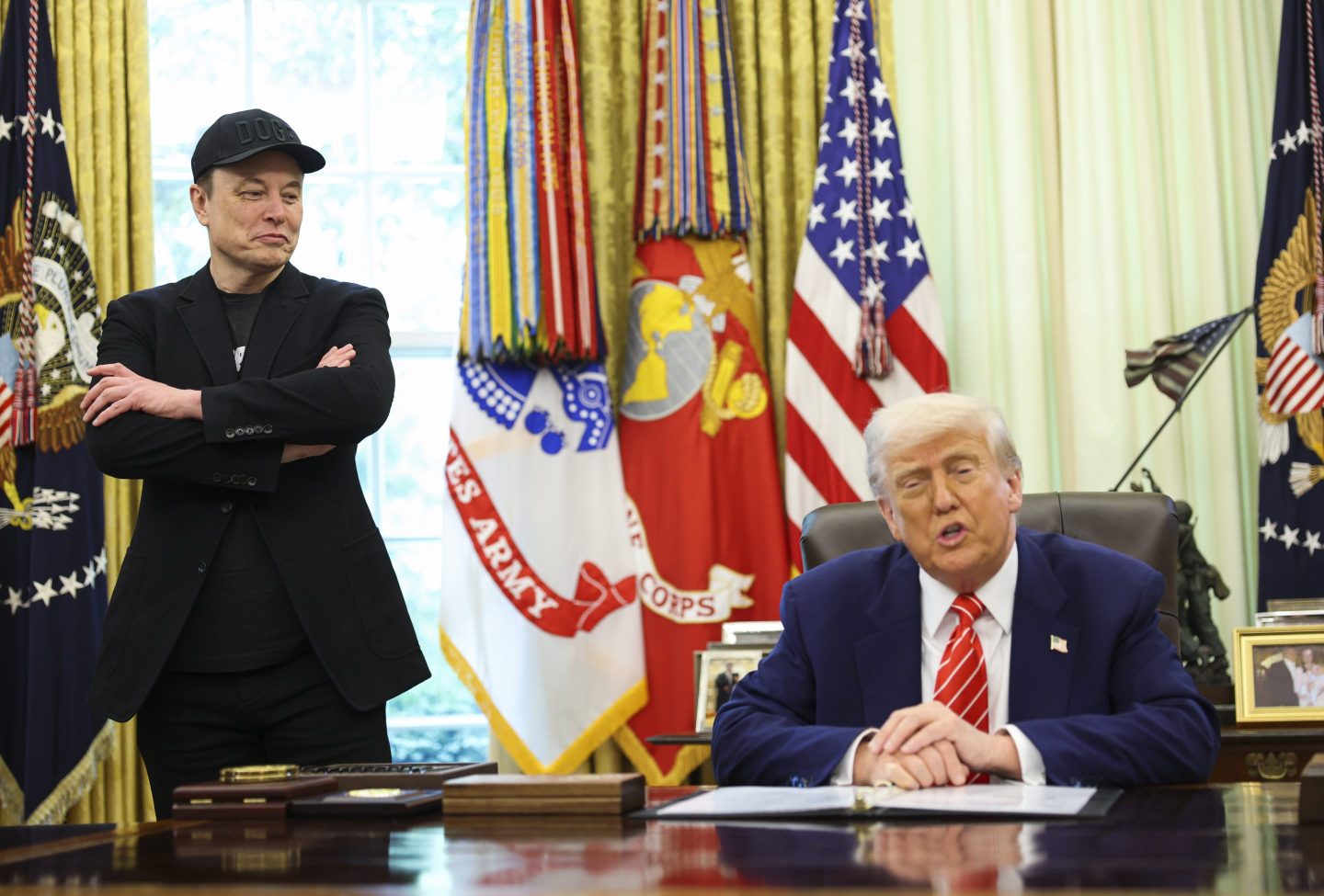 Musk stands with his arms cross next to Trump who sits a table.
