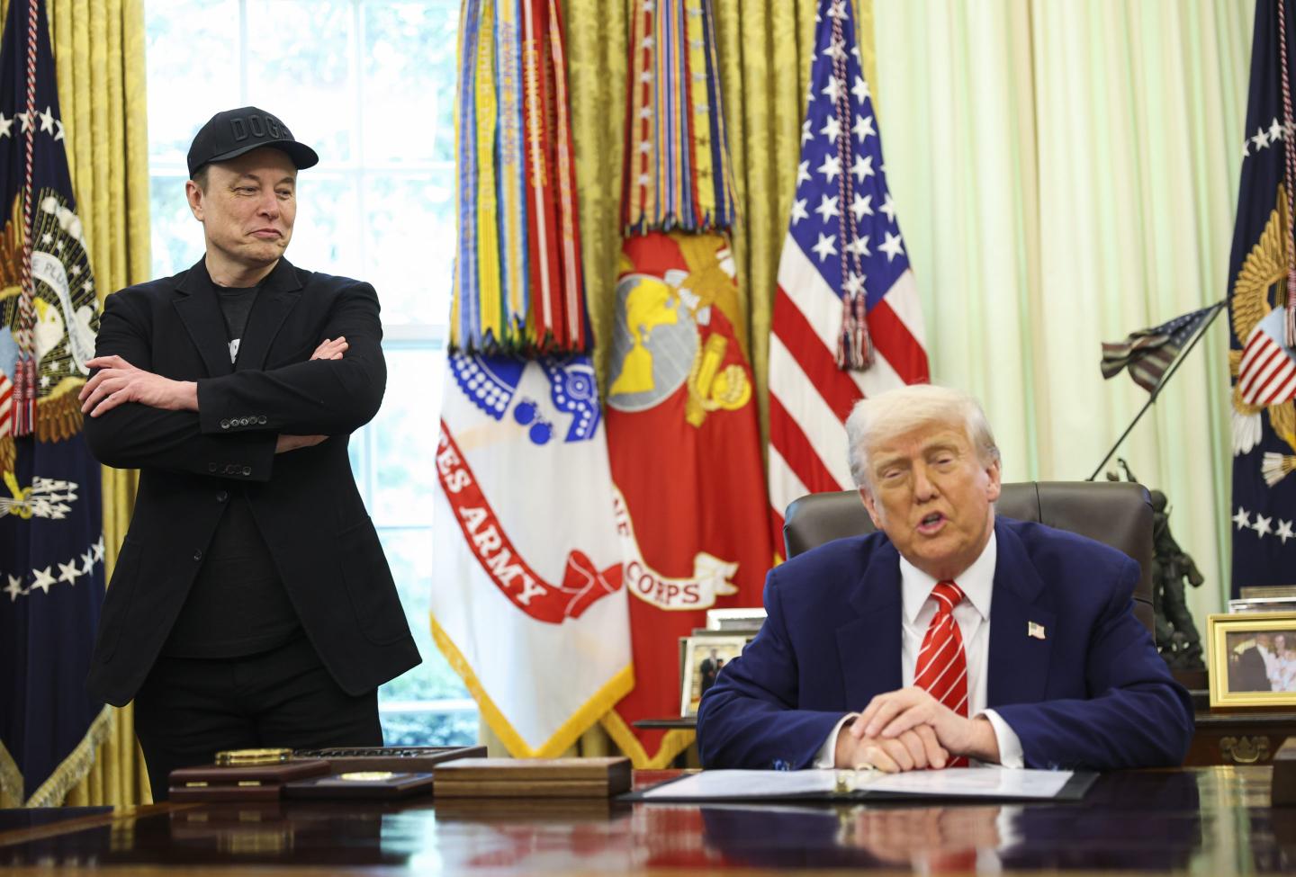 Musk stands with his arms cross next to Trump who sits a table.