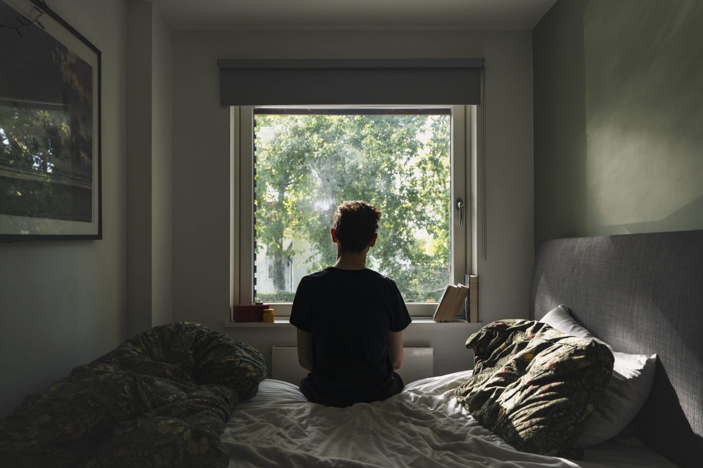 Rear view of a depressed non binary person looking out of their bedroom window on a sunny day - negative emotion
