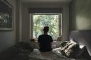 Rear view of a depressed non binary person looking out of their bedroom window on a sunny day - negative emotion