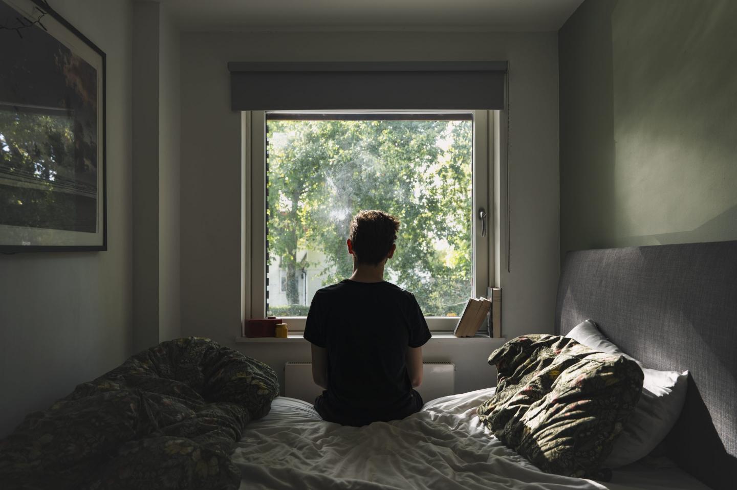 Rear view of a depressed non binary person looking out of their bedroom window on a sunny day - negative emotion