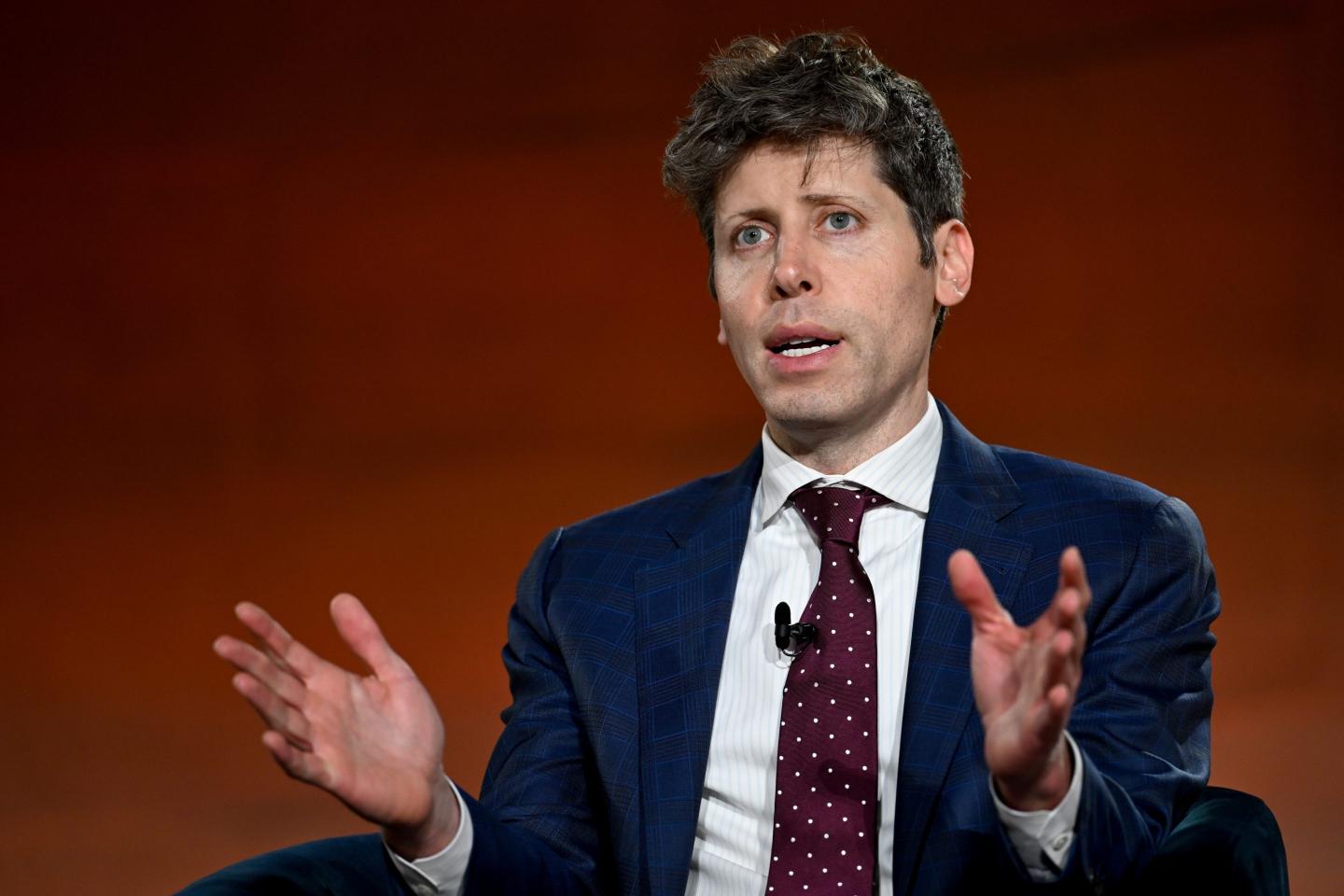 Sam Altman, wearing a suit, speaks in front of a dark red background.