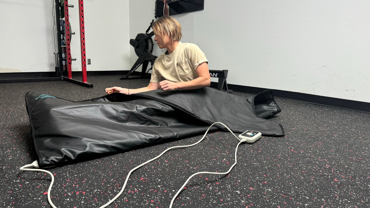 A woman tucked into the HigherDOSE Infrared Sauna Blanket. She's sitting up, and a cord extends from the blanket to the handheld control panel