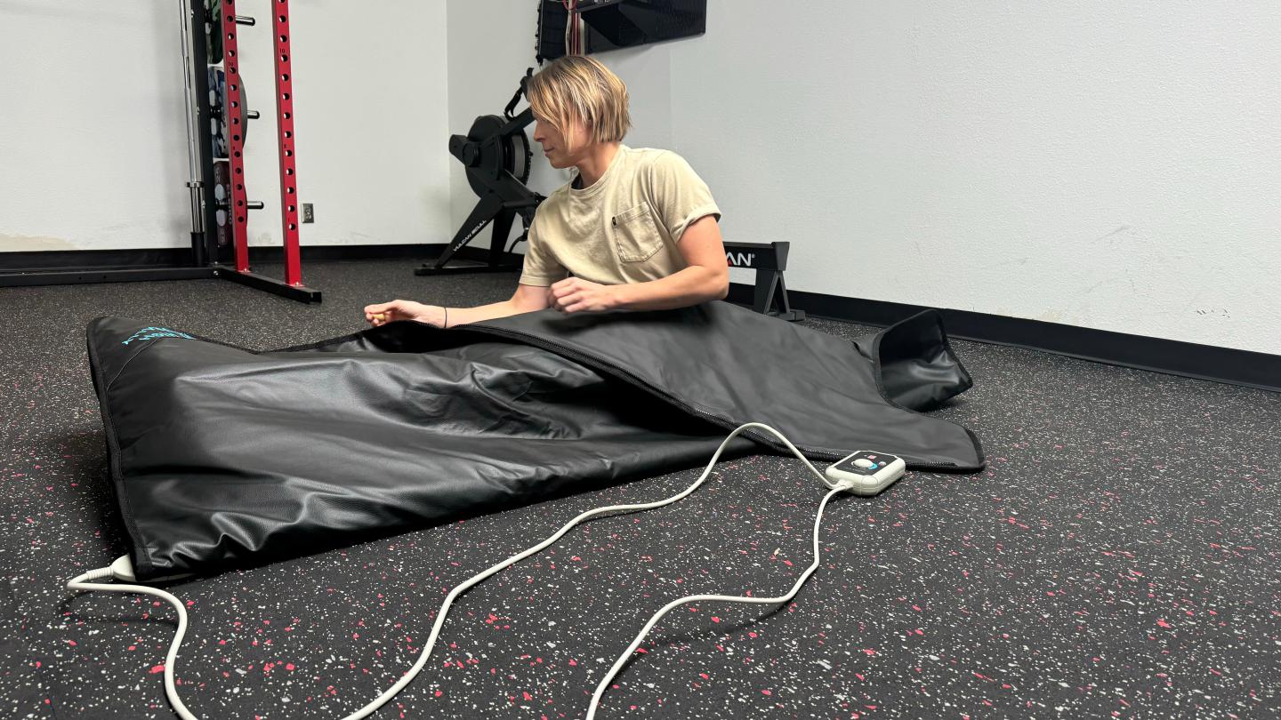 A woman tucked into the HigherDOSE Infrared Sauna Blanket. She's sitting up, and a cord extends from the blanket to the handheld control panel