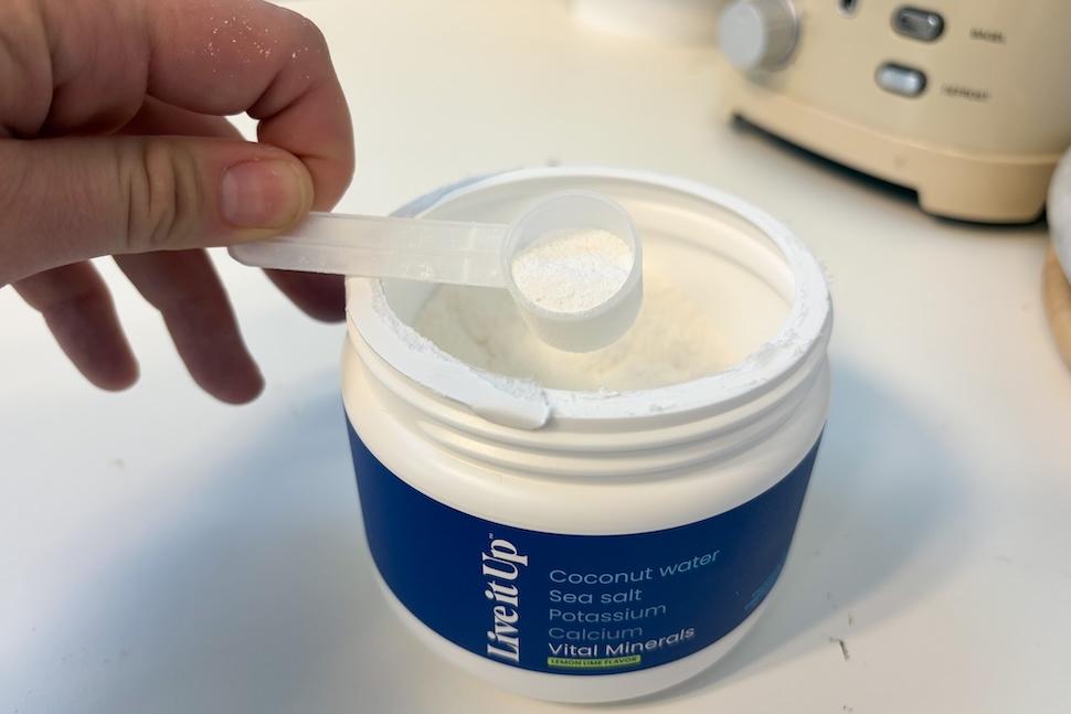 An open canister of Live it Up Vital Minerals. A fine white powder can be seen inside, and a hand is using a plastic scooper to scoop out some of it