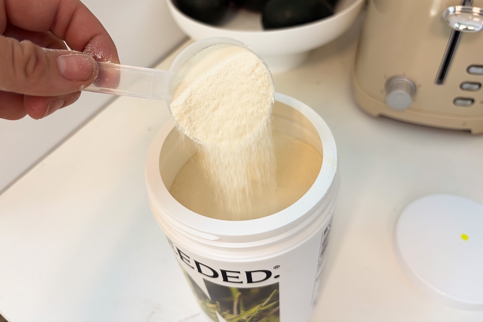 An open canister of Needed Collagen Protein sitting on a white countertop. A hand is using a plastic scooper to take out one scoop of the off-white powder.
