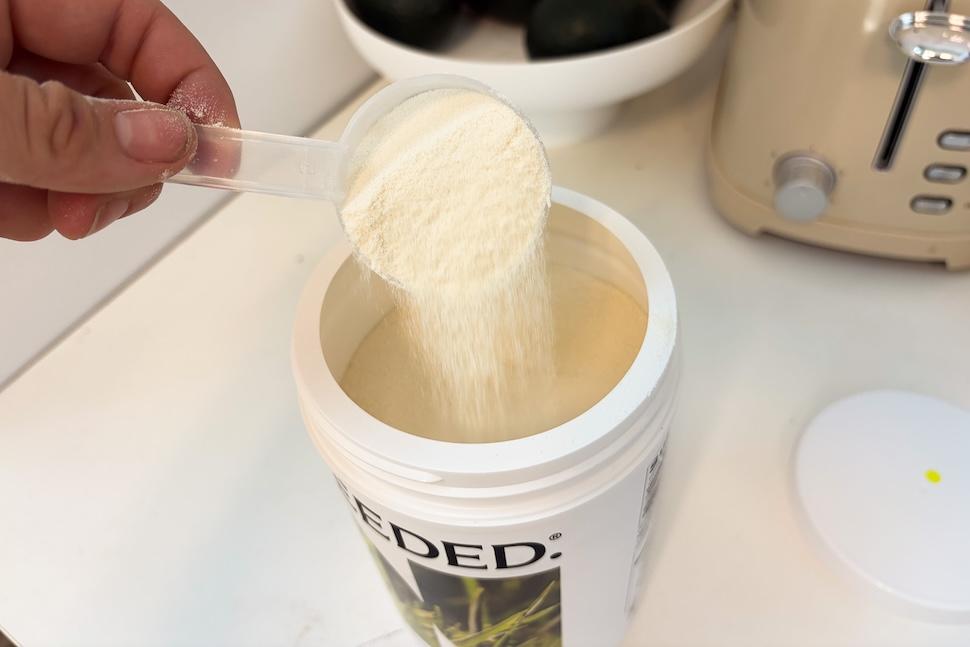 An open canister of Needed Collagen Protein sitting on a white countertop. A hand is using a plastic scooper to take out one scoop of the off-white powder.
