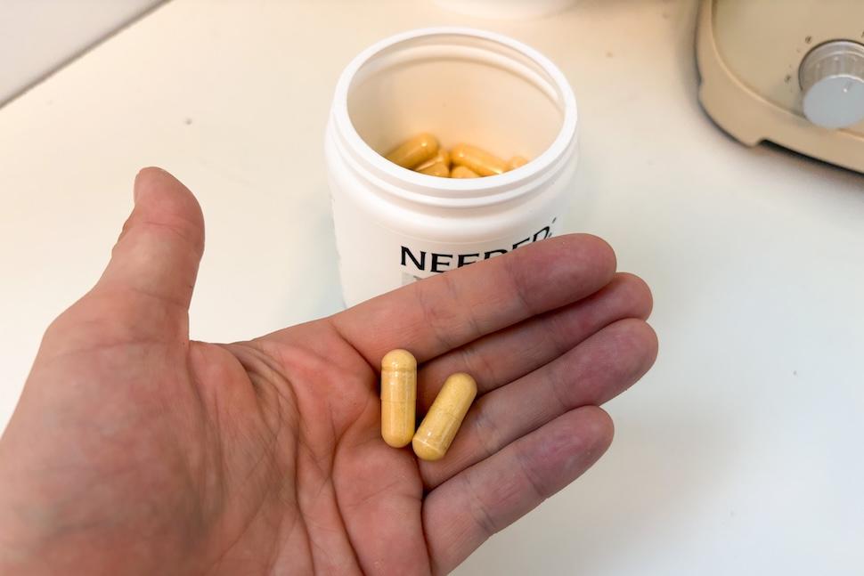 An open palm holding two Needed Men's Multi capsules. The capsules are yellowish in color. Below the hand is an open canister of the capsules on a white countertop.
