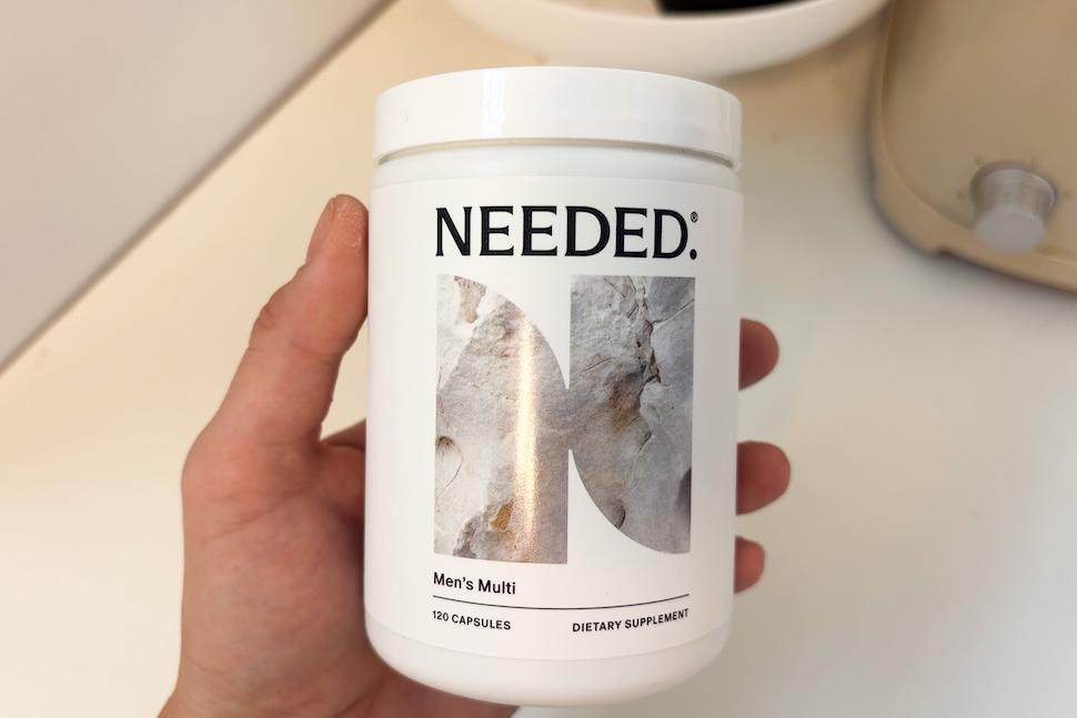 A hand holding a white canister of Needed Men's Multi supplements.