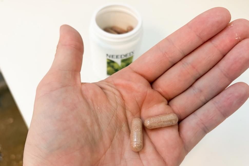 An open palm holding two Needed Pre/Probiotic capsules. On the countertop below, an open canister of the supplements is visible.