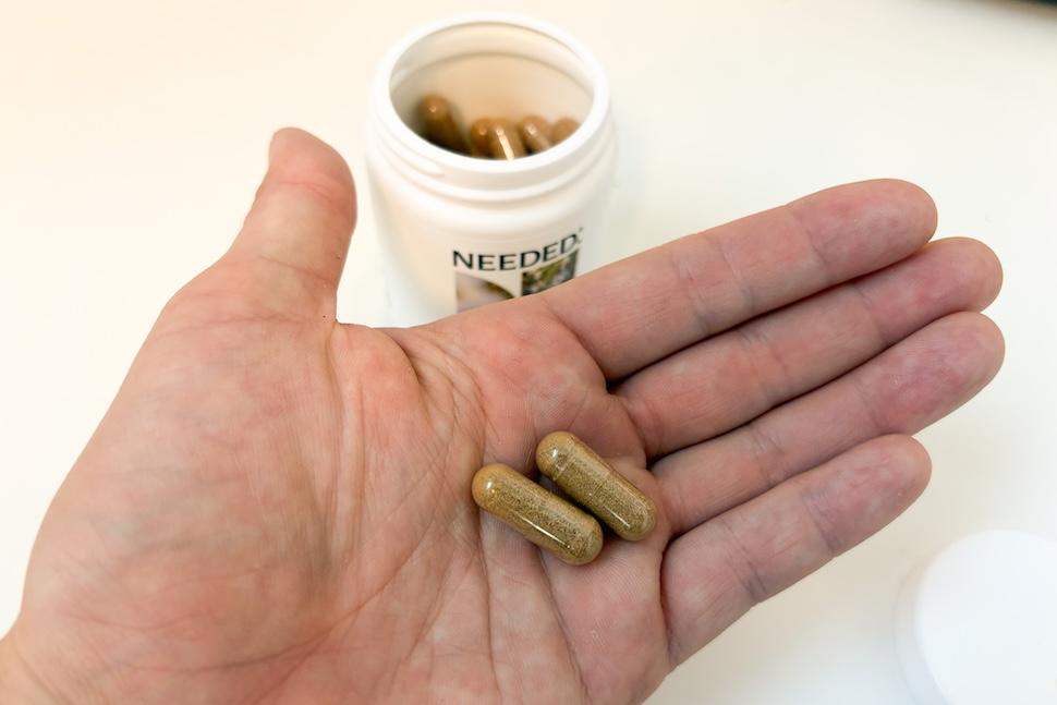 An open palm holding two Needed Stress Support capsules. The capsules of a goldish-brown color to them. Below, an open canister of the capsules can be seen on a white countertop.