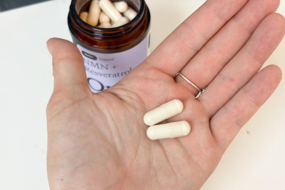 An open palm holding two Omre NMN capsules. They are off-white in color. On the countertop below is an open jar of the capsules.