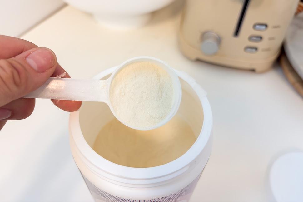 An open canister of Organifi Unflavored Collagen. A hand is scooping out a single scoop of the fine, off-white powder.