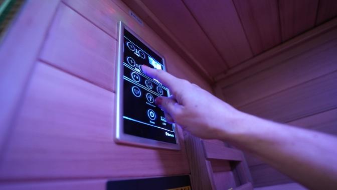 The control panel of the Sun Home Sauna Luminar 5-Person Outdoor Sauna. A woman's hand is reaching up and pressing the touchscreen.