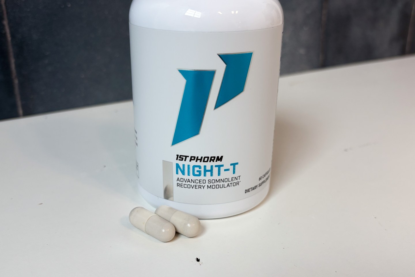 A bottle of 1st Phorm Night-T sitting on a white counter. Two off-white capsules are in front of the bottle.