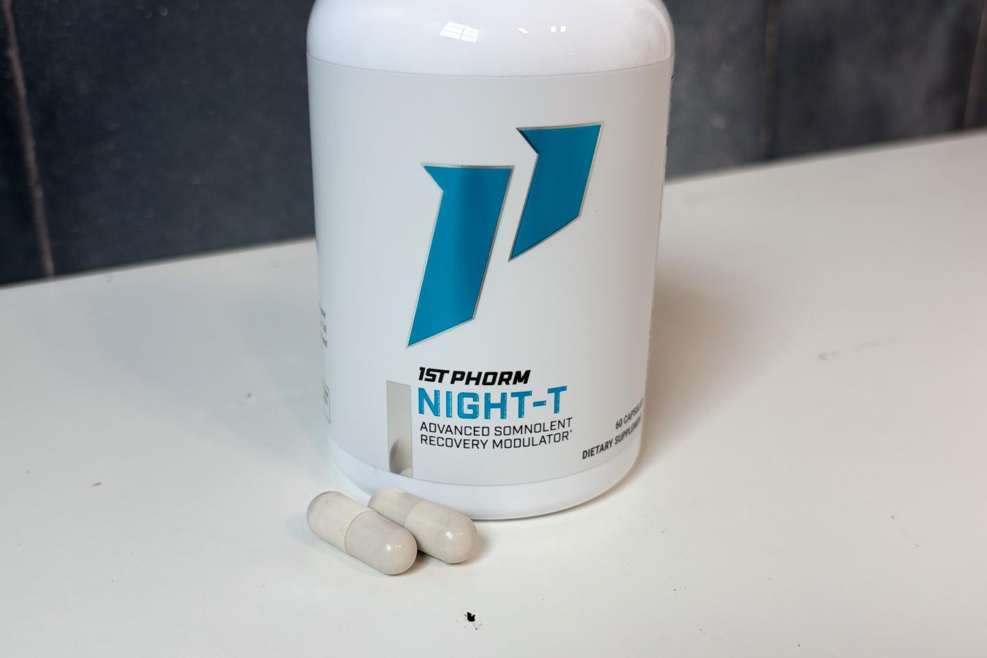 A bottle of 1st Phorm Night-T sitting on a white counter. Two off-white capsules are in front of the bottle.