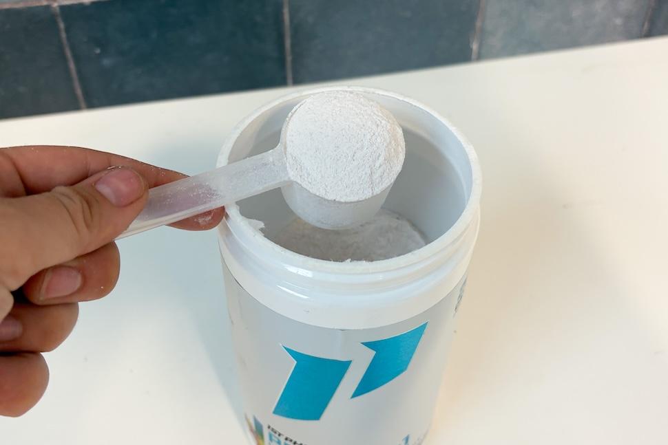 An open canister of 1st Phorm Project-1 sitting on a white countertop. A hand is using a plastic scooper to scoop out the fine white powder.
