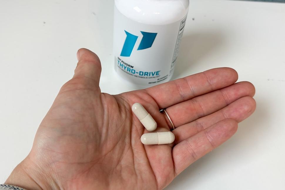 An open palm holding two 1st Phorm Thyro Drive capsules, which are off-white in color. A bottle of the capsules sits nest to the hand on a white countertop.