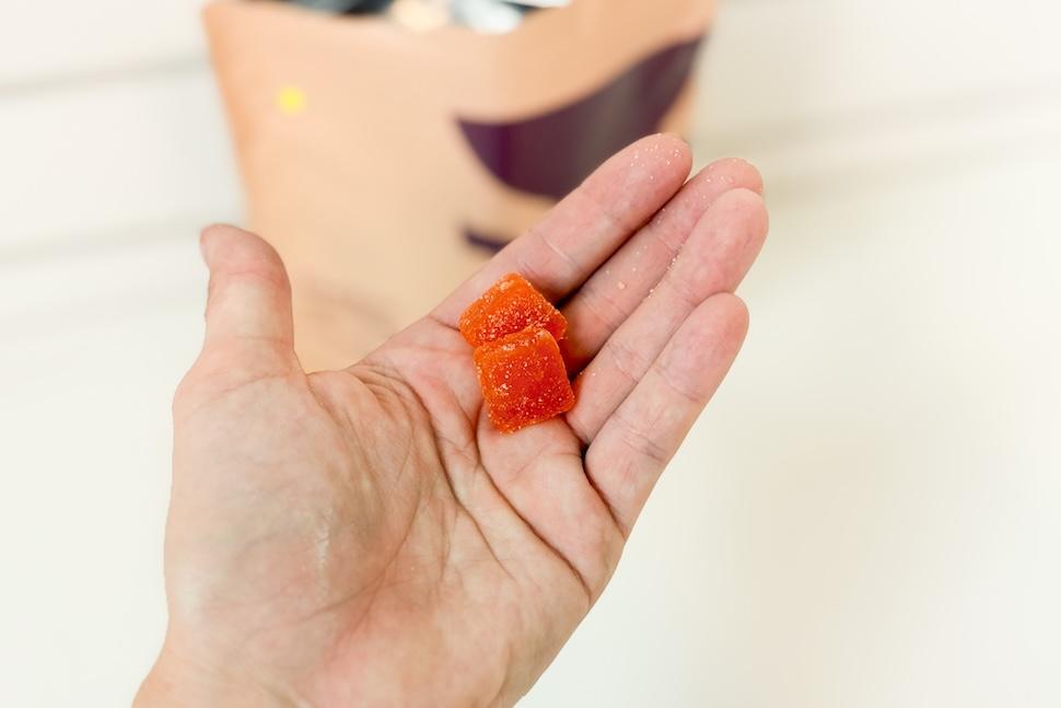 An open palm holding two orange Bird&Be Gentle Prenatal Gummies. You can see the tan bag of gummies on the white countertop below.