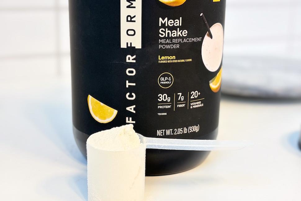 A canister of Facto Form Meal Replacement sitting on a countertop. A plastic scoop sits next to it, packed to the brim with off-white powder.