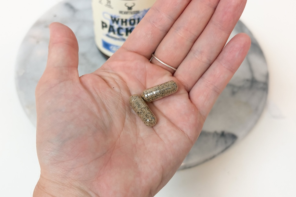 An open palm holding two Heart & Soil Whole Package capsules. The capsules are see-through, with black and gold flecks inside.