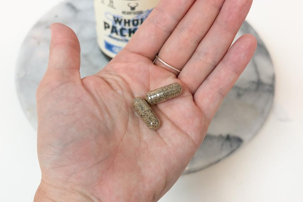 An open palm holding two Heart & Soil Whole Package capsules. The capsules are see-through, with black and gold flecks inside.
