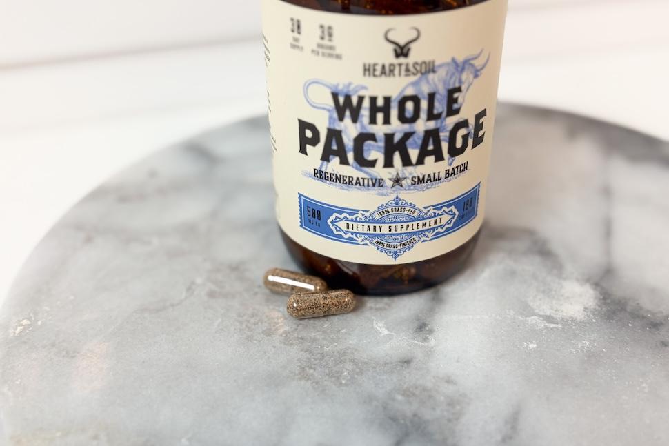 A bottle of Heart & Soil Whole Package. Two golden-hued pills sit next to it on a marble countertop.