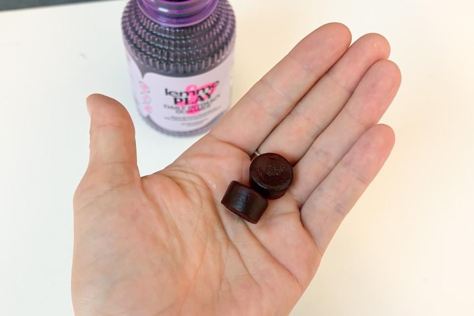 An open palm holding two Lemme Play gummies. The gummies are ruby red in color. On the countertop below, you can see the purple bottle of gummies.
