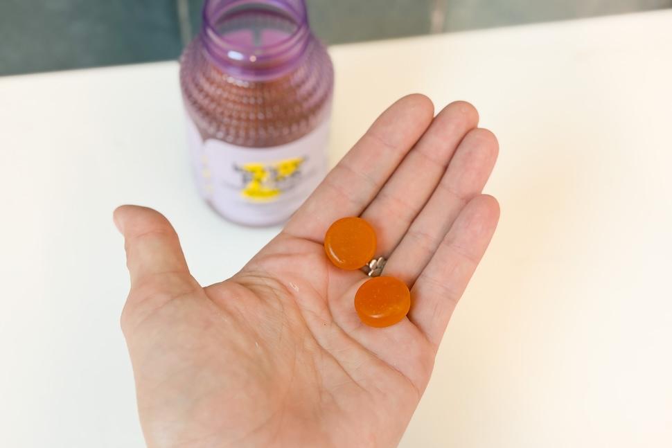 An open palm holding two orange Lemme Purr gummies. Below, you can see the open bottle of gummies on an off-white countertop.