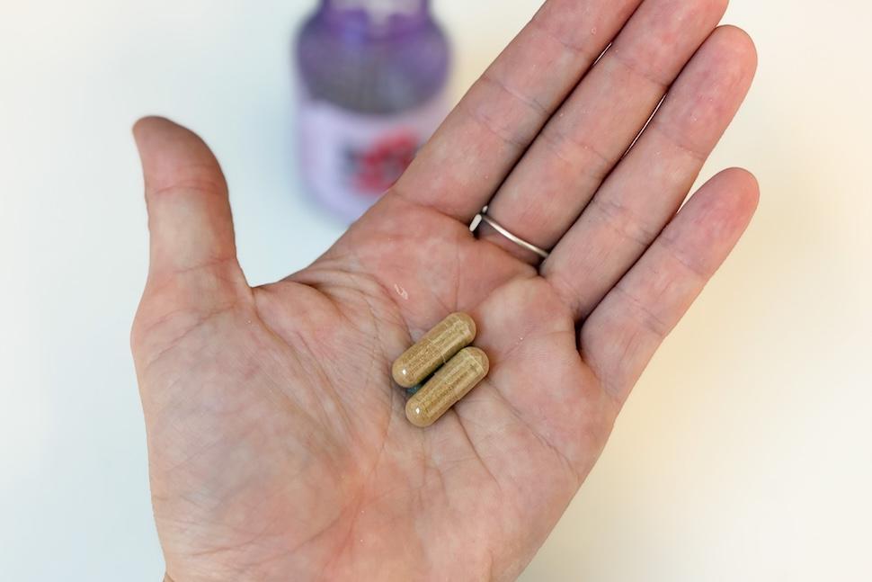 An open palm holding two Lemme Burn capsules. The capsules are a gold-ish color. One the white countertop below, you can see the purple bottle of the supplements.