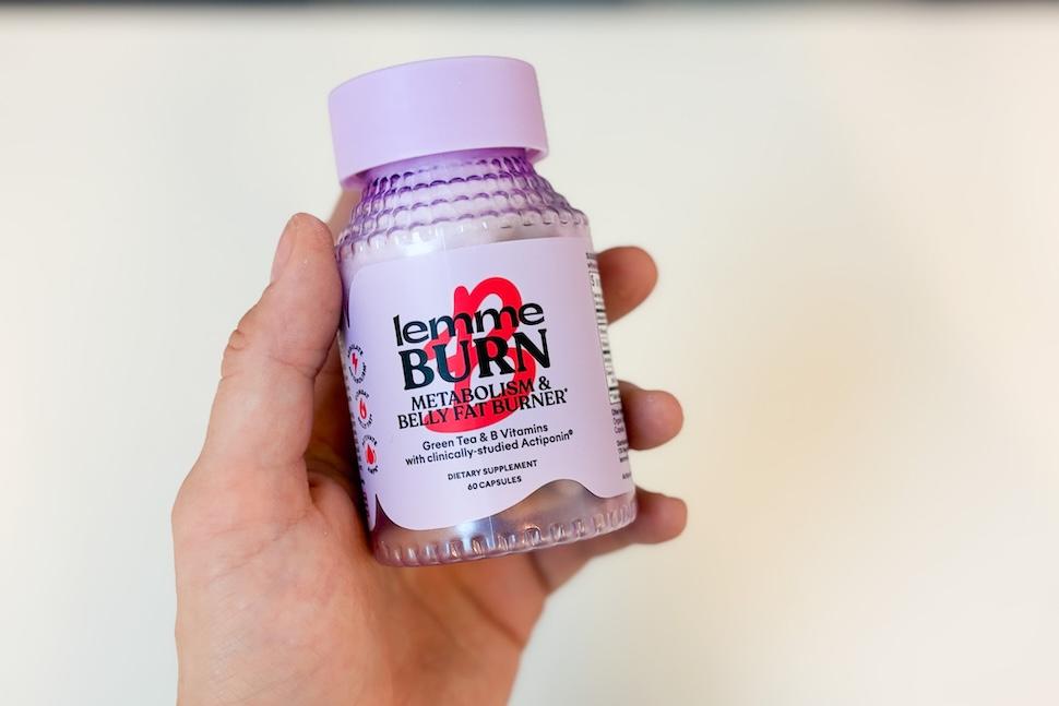 Closeup of a hand holding a bottle of Lemme Burn supplements. The bottle has a purple tint to it.
