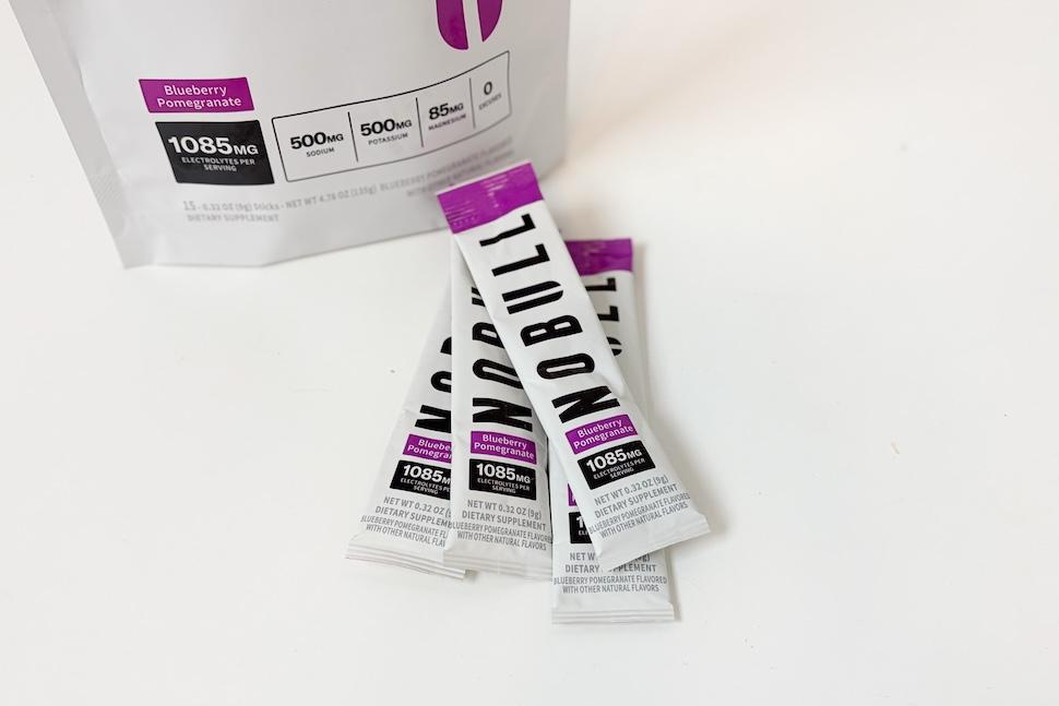 Several packets of NOBULL Electrolytes lying on a white countertop. A hand holding a packet of NOBULL Electrolytes. The packaging is white and purple and the flavor if blueberry pomegranate.