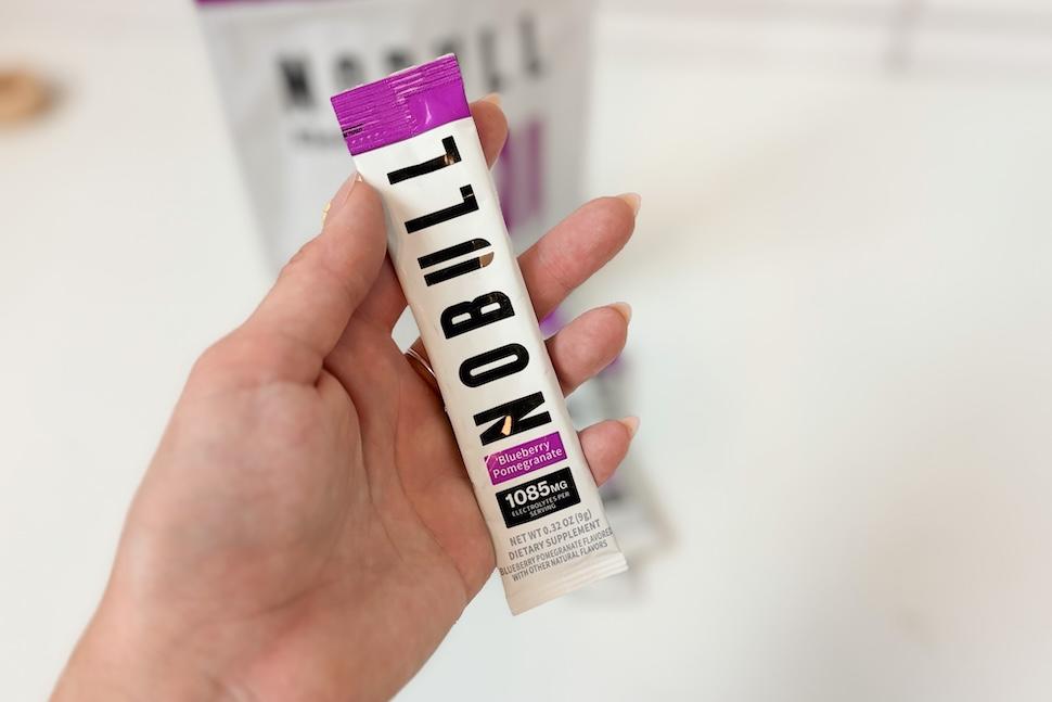 A hand holding a packet of NOBULL Electrolytes. The packaging is white and purple and the flavor if blueberry pomegranate.