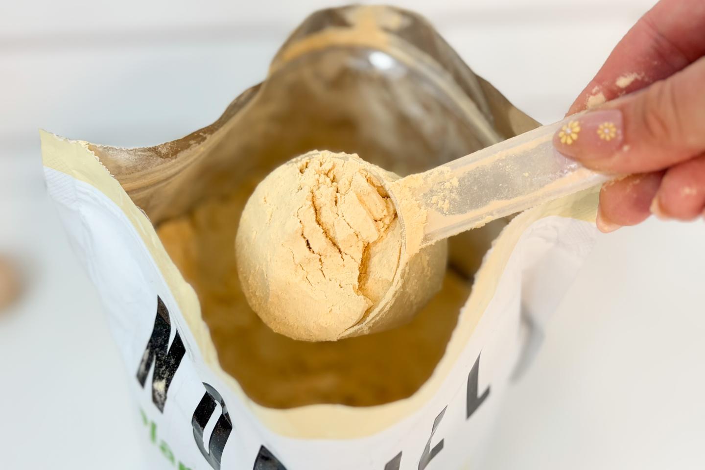 An open bag of NOBULL Plant Protein. A hand is scooping out the off-white powder with a plastic scoop.