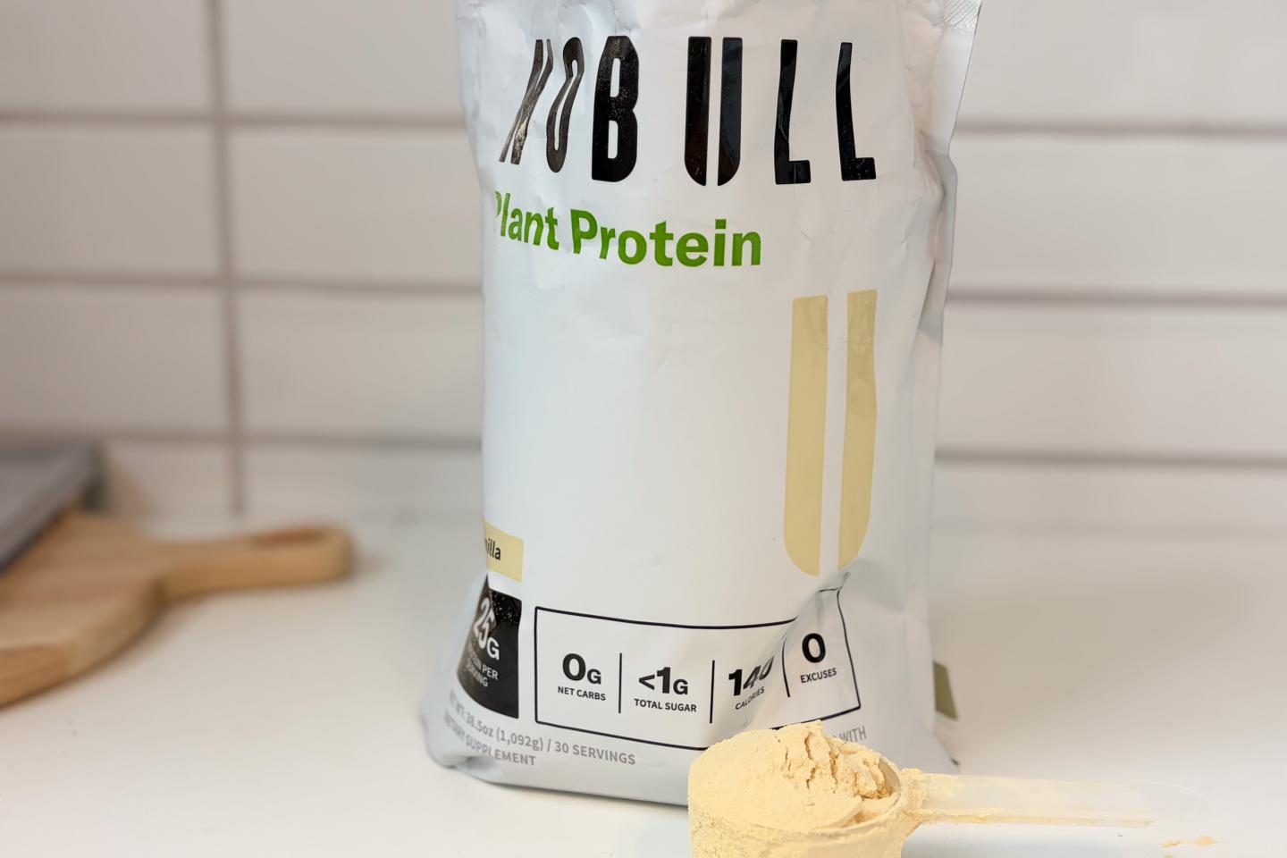 A bag of NOBULL Plant Protein sitting on a white countertop. A scoop packed with the off-white powder sits next to it.