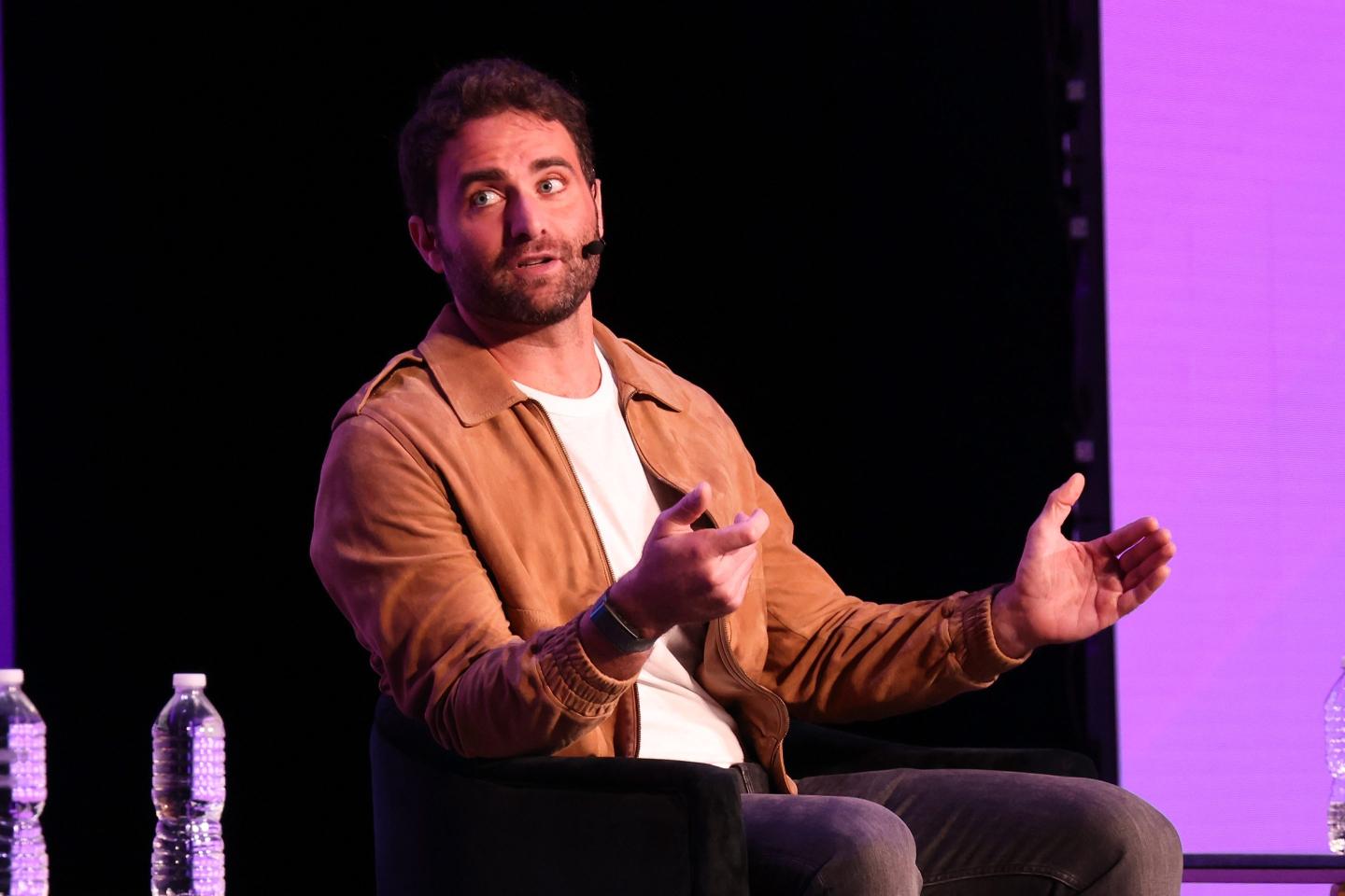 Peter Rahal speaks on stage in front of a black and purple background.