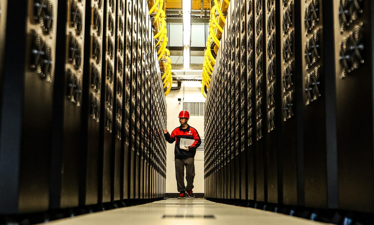 A man wearing a red and black jacket and a red hat walks down a hallway lined with servers.