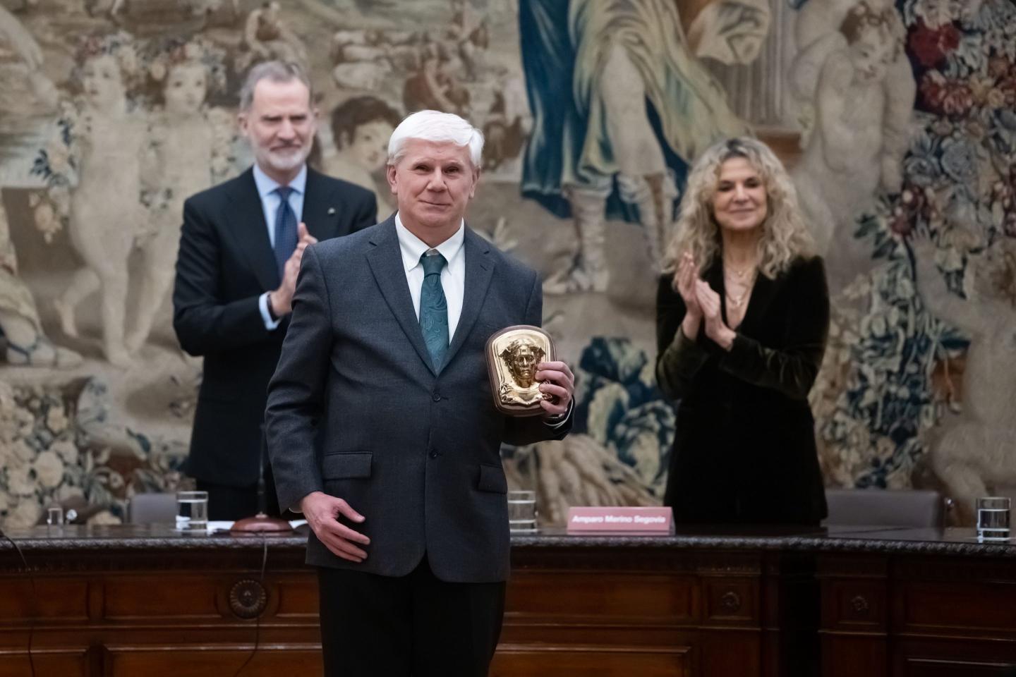 Brown University Professor Roberto Serrano, a man in a suit holding onto a gold trophy--the King Of Spain Economy Award"-- before Spain's King Felipe and a painted wall.