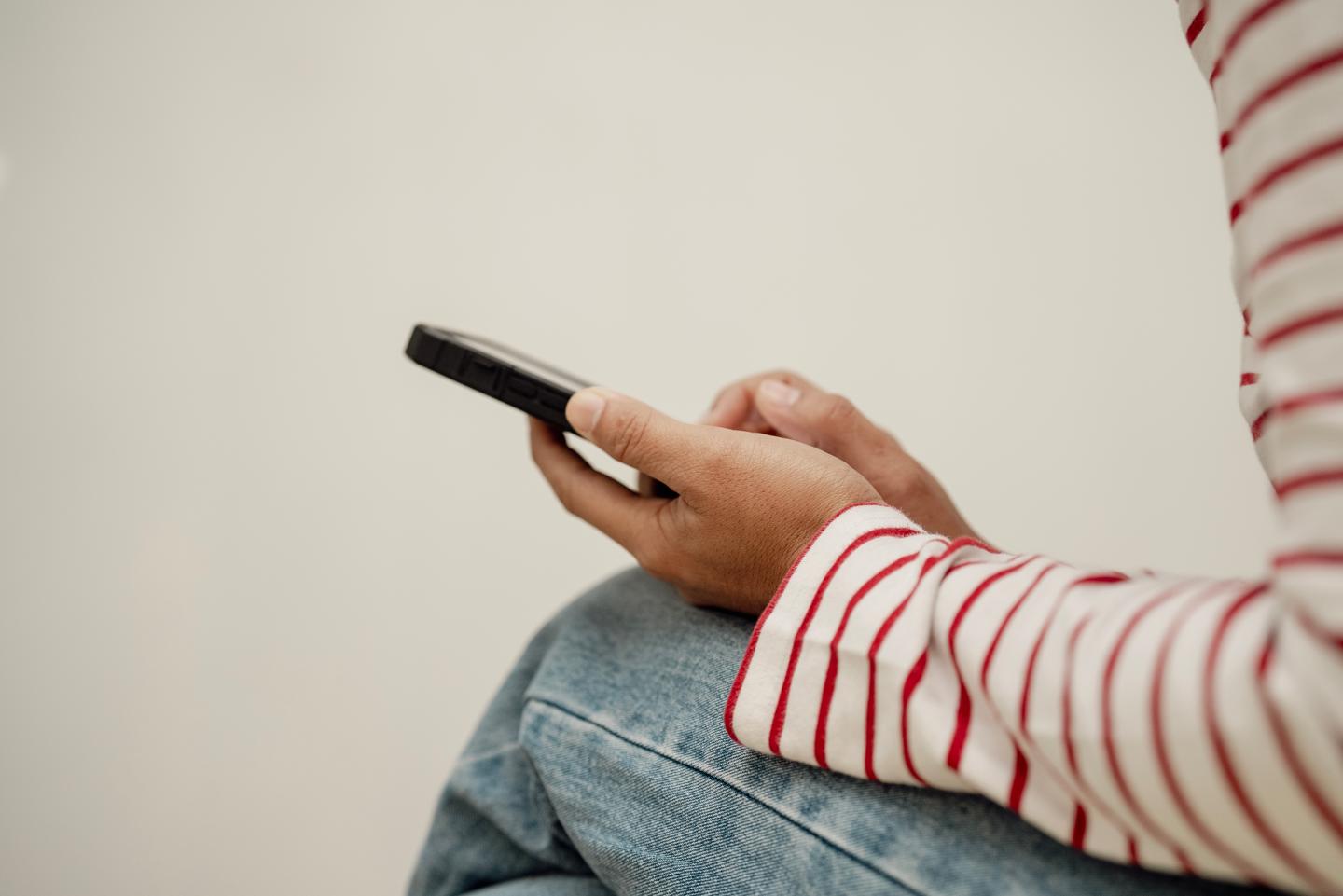 A close-up view of a woman wearing a striped shirt and jeans, sitting and using a smartphone with one hand in a casual setting.