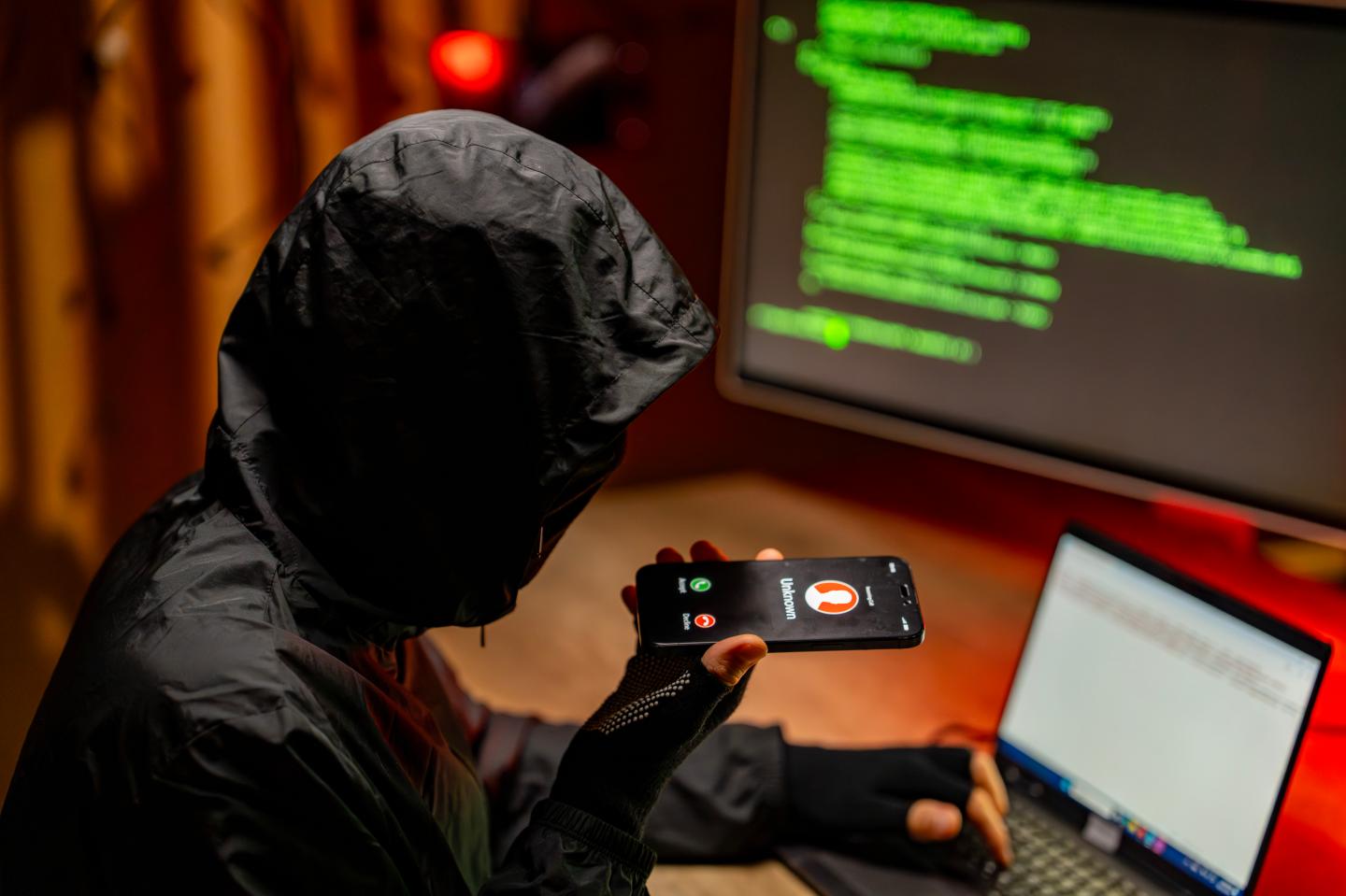 Hacker in a dark hooded jacket holding a smartphone displaying an incoming unknown call while working on a laptop
