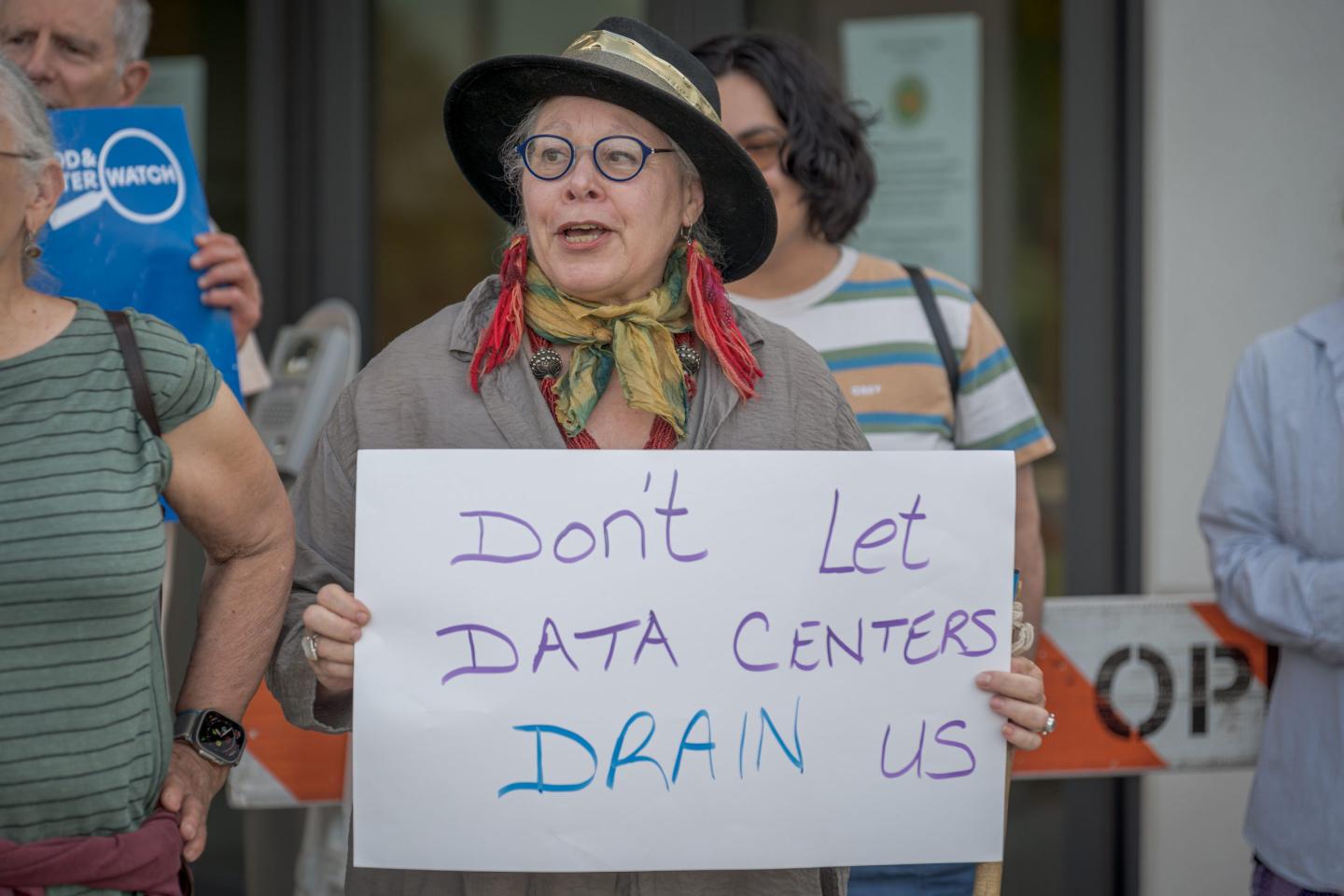 A woman wearing a hat and scarf holds a sign that says, "Don't let data centers drain us."