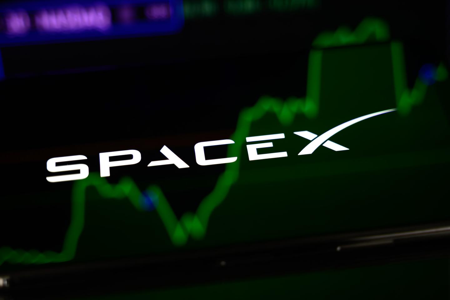 The SpaceX logo is displayed on the screen of a smartphone placed on a reflective surface onto which a stock market chart is projected.