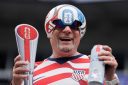 A man in a soccer ball hat holds a trophy and a beer in his hands.