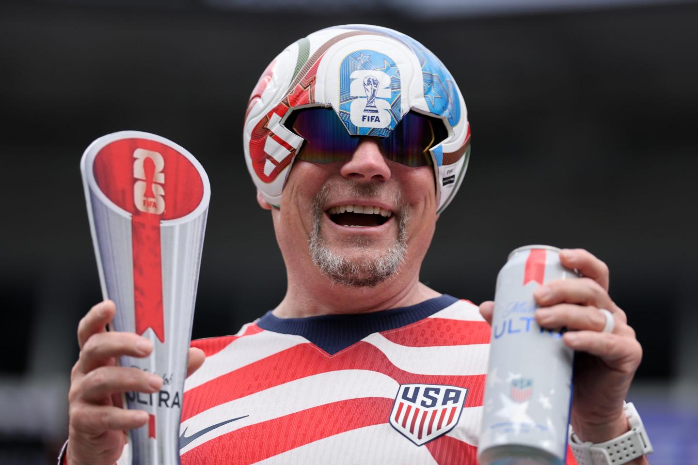 A man in a soccer ball hat holds a trophy and a beer in his hands.
