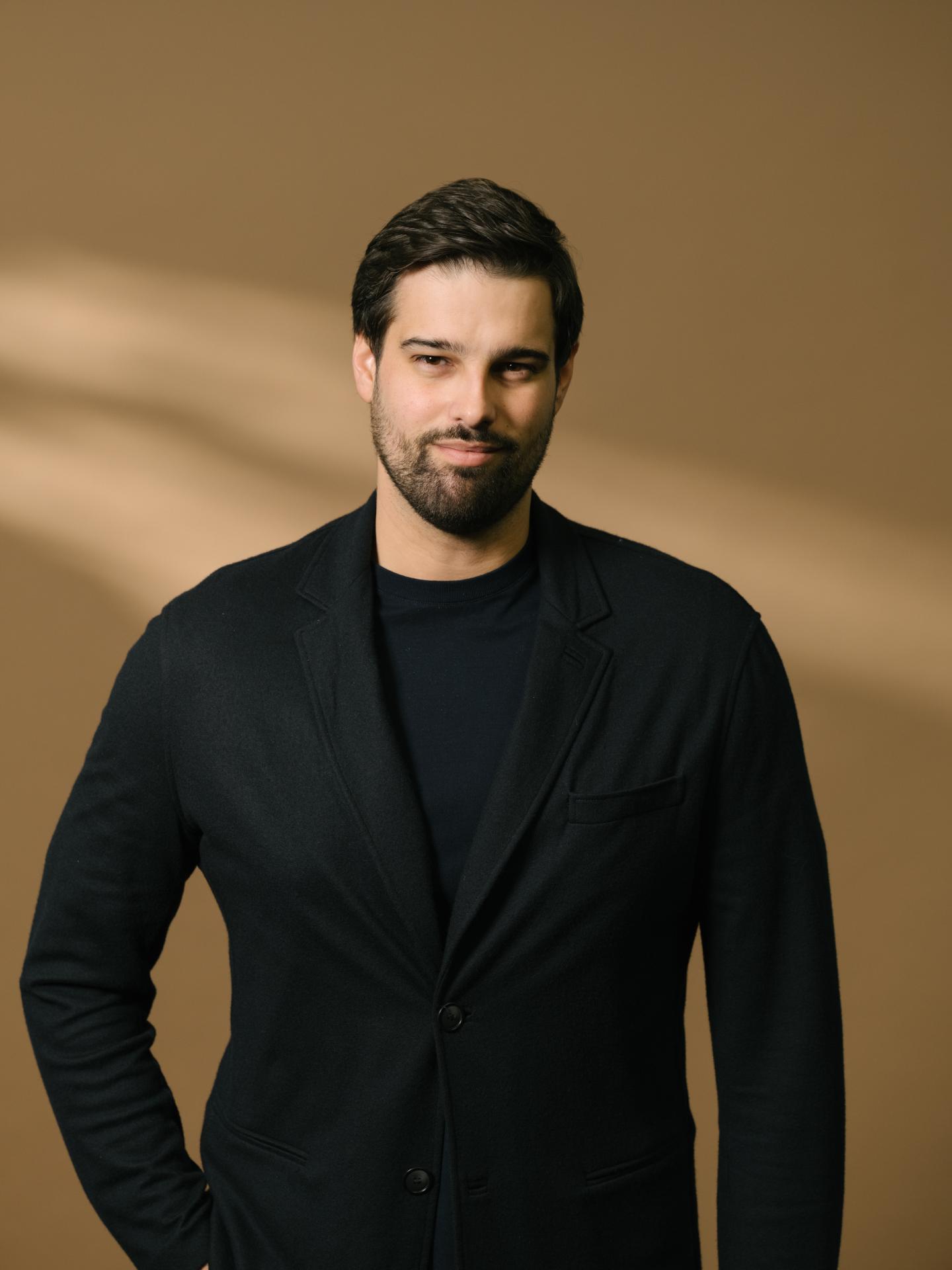 Microsoft executive Jacob Andreou poses for a photo wearing a black suit jacket, with a tan background behind him.