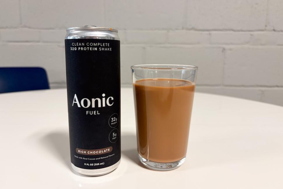 A black can of Aonic Fuel protein shake sits next to a glass full of the liquid on a white countertop.
