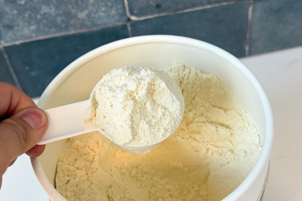 The inside of a canister of Heart & Soil Grass-Fed Whey Protein. A hand is using a plastic scoop to scoop out the off-white powder.