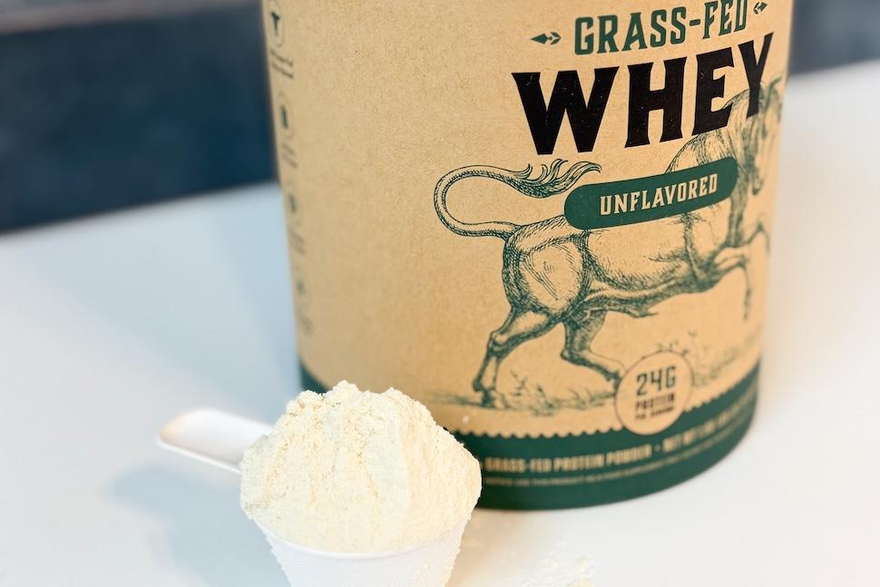 A canister of Heart & Soil Grass-Fed Whey Protein. A plastic scoop packed with the off-white powder sits next to it on the counter.