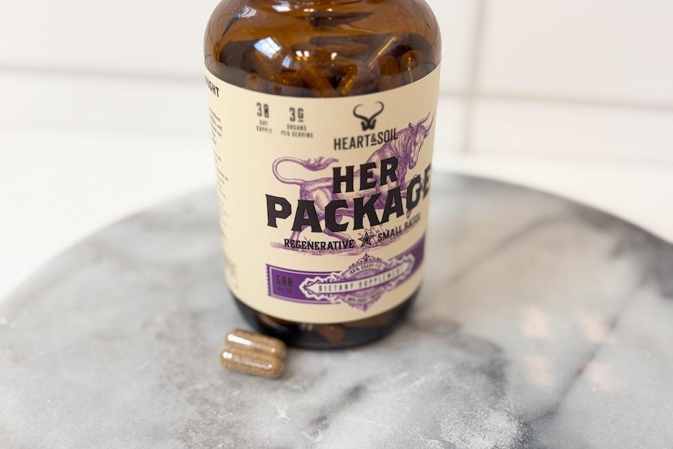 A bottle of Heart & Soil Her Package sitting on a marble countertop. Two golden-hued pills sit next to the bottle.