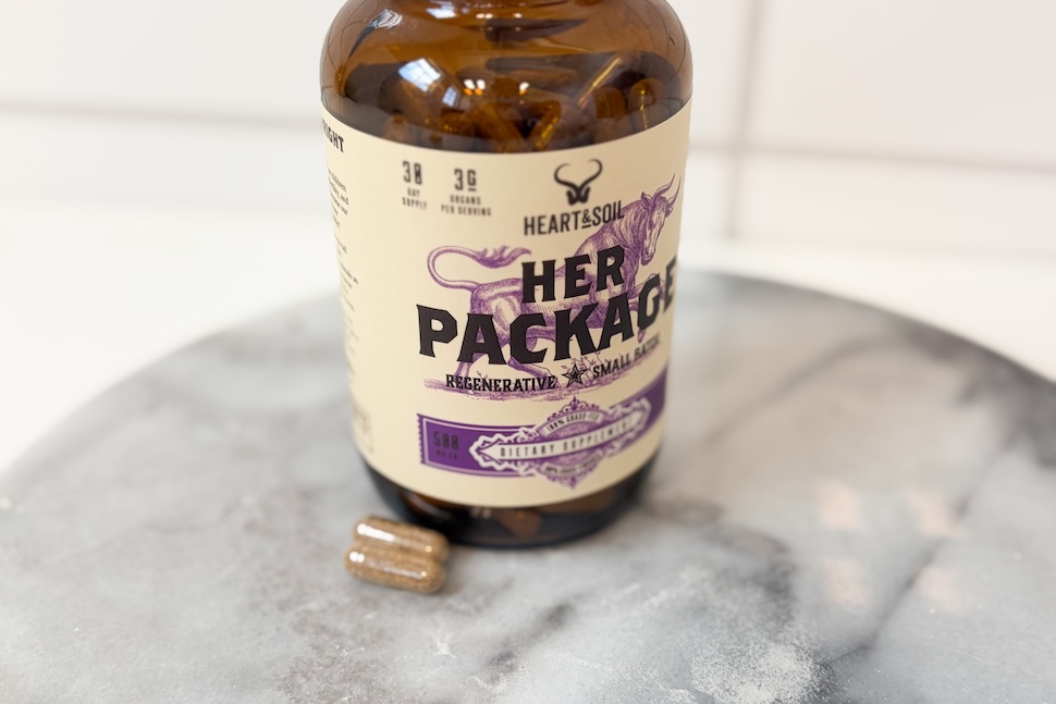 A bottle of Heart & Soil Her Package sitting on a marble countertop. Two golden-hued pills sit next to the bottle.